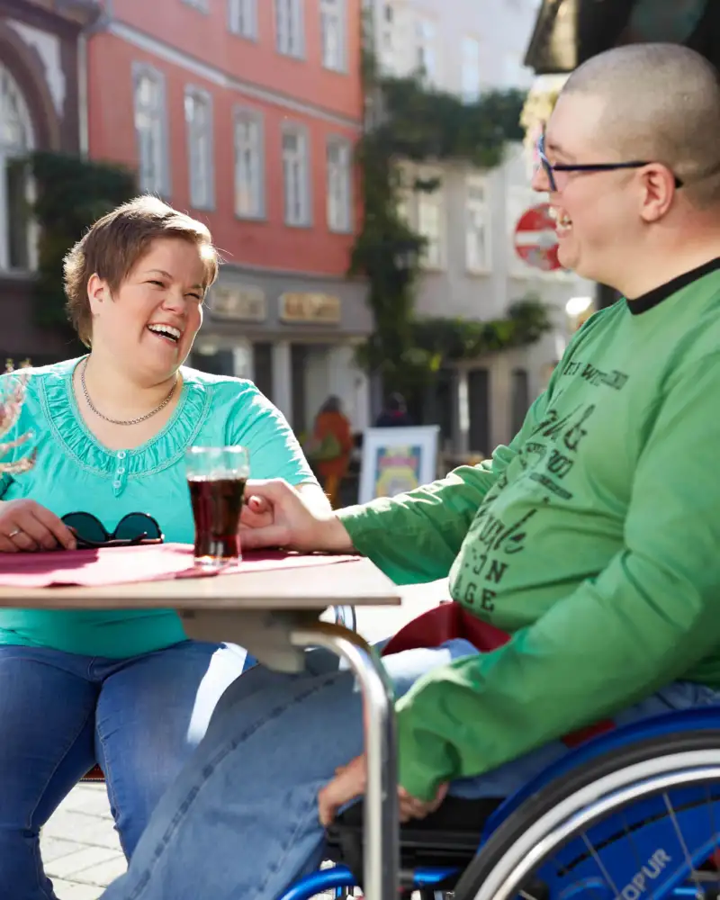Eine Frau und ein Mann im Rollstuhl sitzen an einem Café-Tisch im Freien