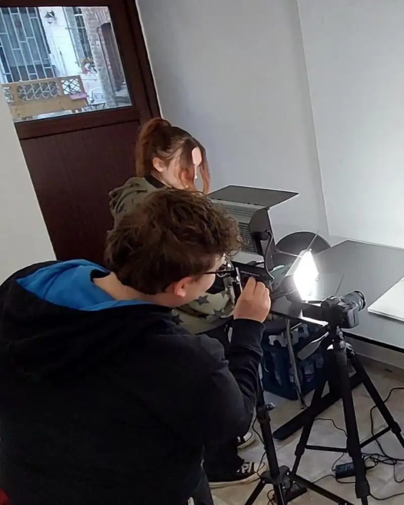 Zwei Frauen und ein Mann schauen auf einen Tisch. Eine Frau schaut durch eine Kamera.