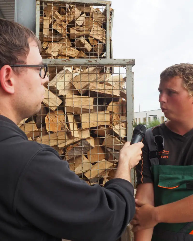 Vor einem Käfig mit Holzstücken stehen zwei junge Männer. Einer trägt eine Arbeitslatzhose. Der andere hält ihm ein Mikrofon hin.