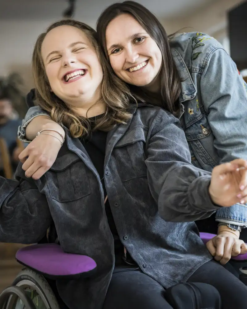 Zwei junge Frauen lachend Arm in Arm, eine von beiden im Rollstuhl