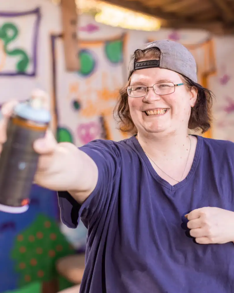 Eine Frau im mittleren Alter steht vor bunt besprühten Wänden. Sie hat eine Sprühdose in der Hand und lacht.