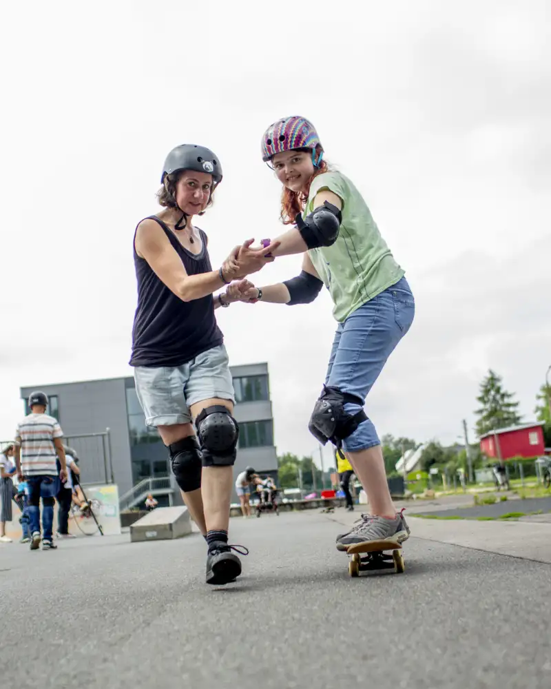 Zwei junge Frauen auf Skateboards. Dabei halten sie sich an beiden Händen fest.
