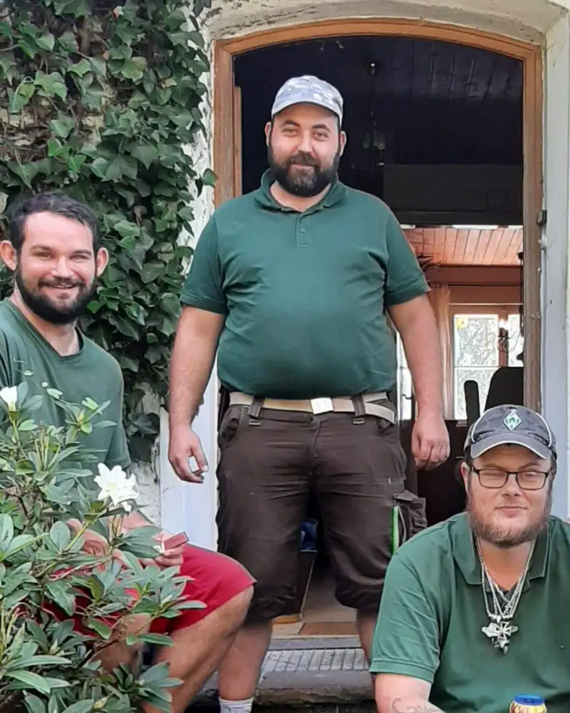 Drei Männer stehen und sitzen auf einer Treppe vor einem Haus. Alle tragen die gleichen grünen T-Shirts.