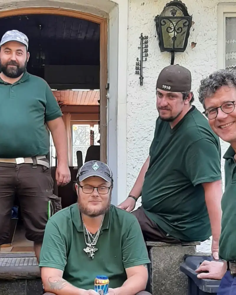Eine Gruppe von Männern stehen und sitzen auf einer Treppe vor einem Haus mit geöffneter Haustür. Alle tragen die gleichen grünen T-Shirts.