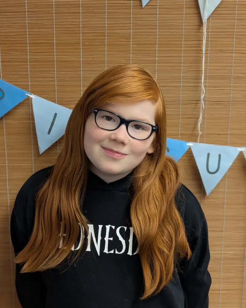 Ein Mädchen mit roten langen Haaren und Brille lächelt in die Kamera.