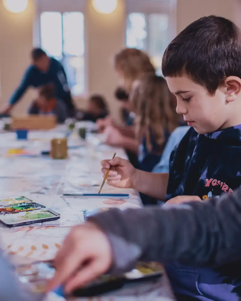 Ein kleiner Junge sitzt mit vielen anderen Kindern an einem langen Tisch und malt.