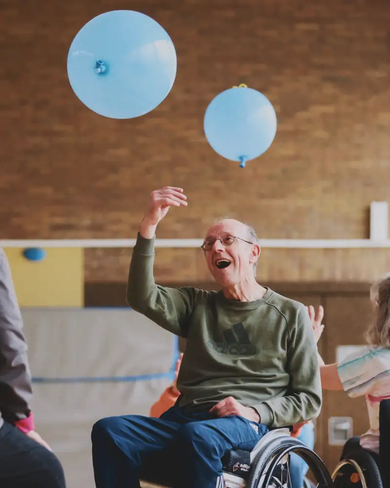 Ein Mann im Rollstuhl stößt mit seiner rechten Hand einen blauen Ballon in die Luft.