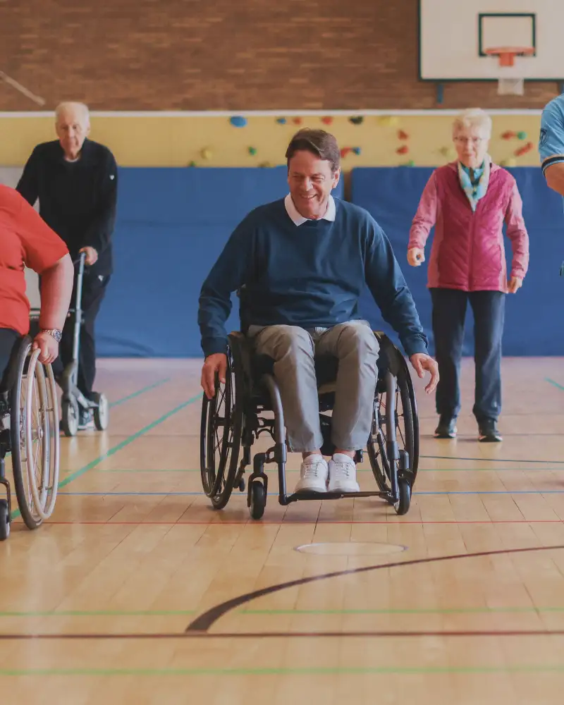 Rudi Cerne und eine Frau sitzen beide in einem Rollstuhl und fahren durch eine Sporthalle.