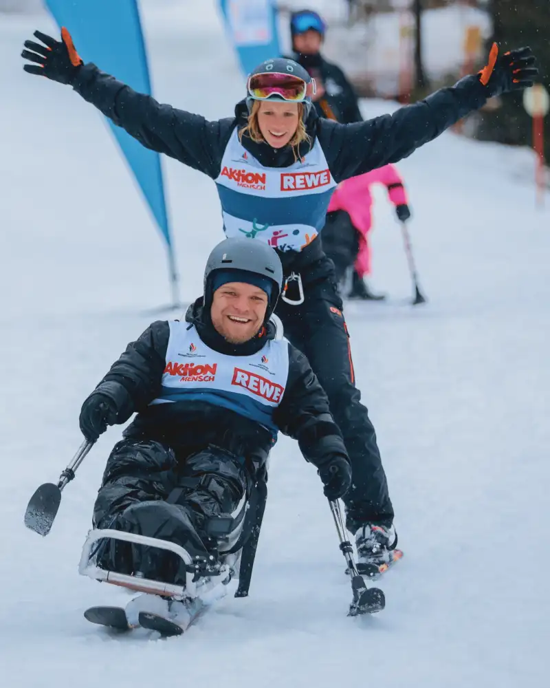 Eine Aufnahme vom Monoski-Event auf der Götschenalm bei Berchtesgaden: Aktion Mensch-Botschafter Niko Kappel hat großen Spaß bei der Abfahrt im Duoski. Hinter ihm reckt eine Begleiterin ihre Arme vor Freude in die Höhe.