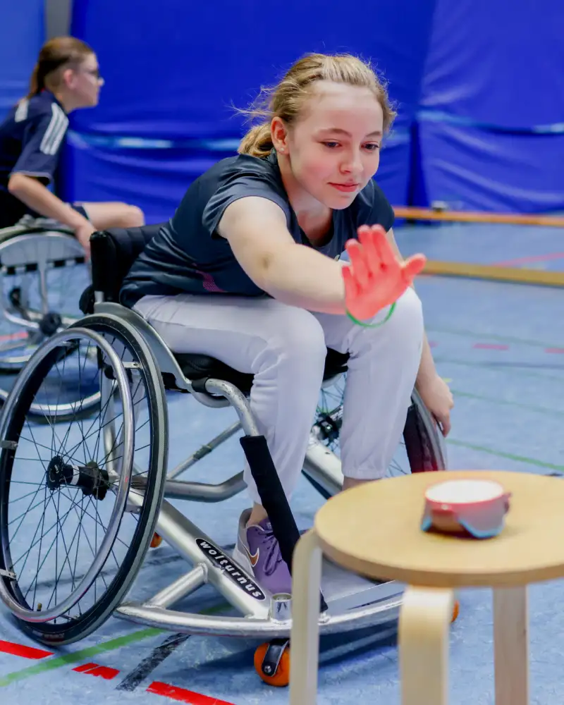 Das Inklusionsmobil auf Schulbesuch: Die Kinder erfahren beim Sport den Umgang mit Rollstuhl, Prothesen und Blindheit. Hier fährt eine Schülerin in einem Sportrollstuhl auf einen Hocker zu, auf dem ein Buzzer liegt.