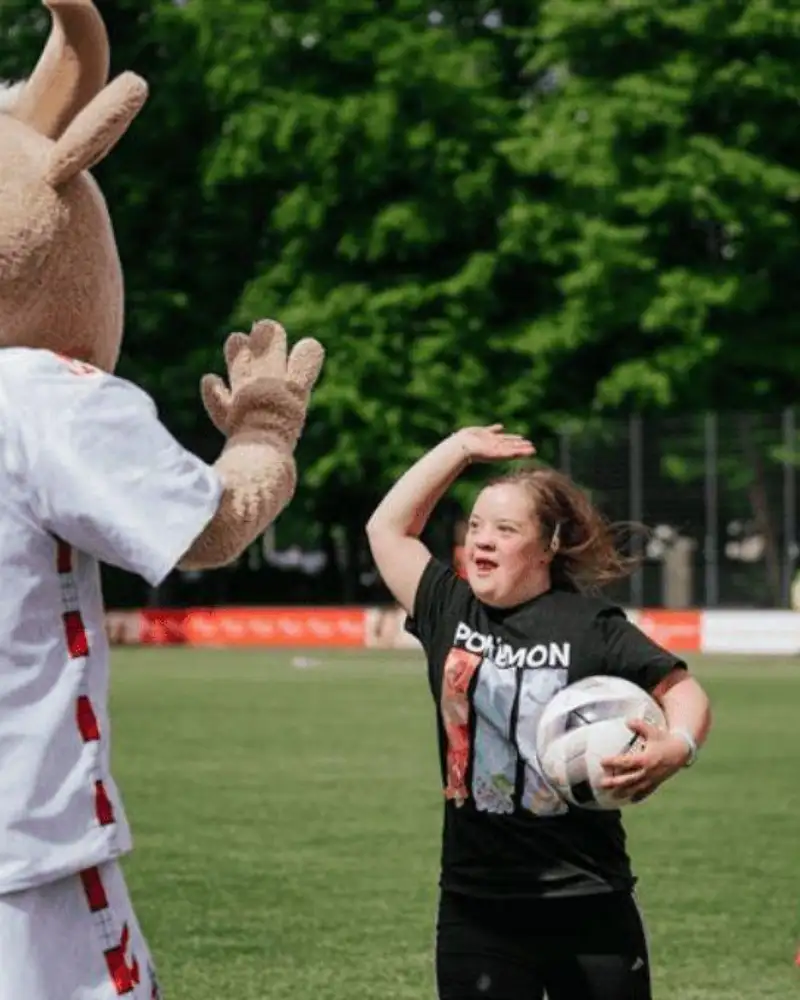 Ein Mädchen mit Down-Syndrom und das Maskottchen des 1. FC Köln geben sich auf dem Fußballfeld ein Highfive.
