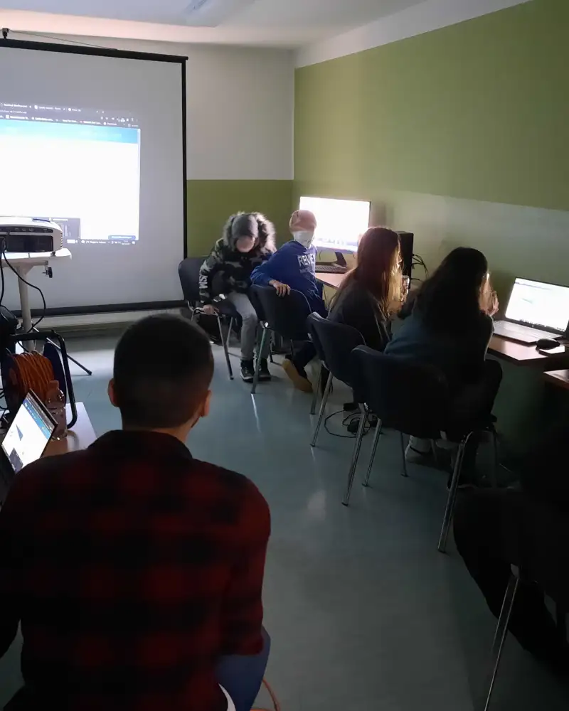 Eine Gruppe von Jugendlichen sitzt in einem abgedunkelten Raum vor leuchtenden Bildschirmen. In der Mitte des Raumes wird ein Bildschirm in Großformat auf eine Leinwand projiziert.