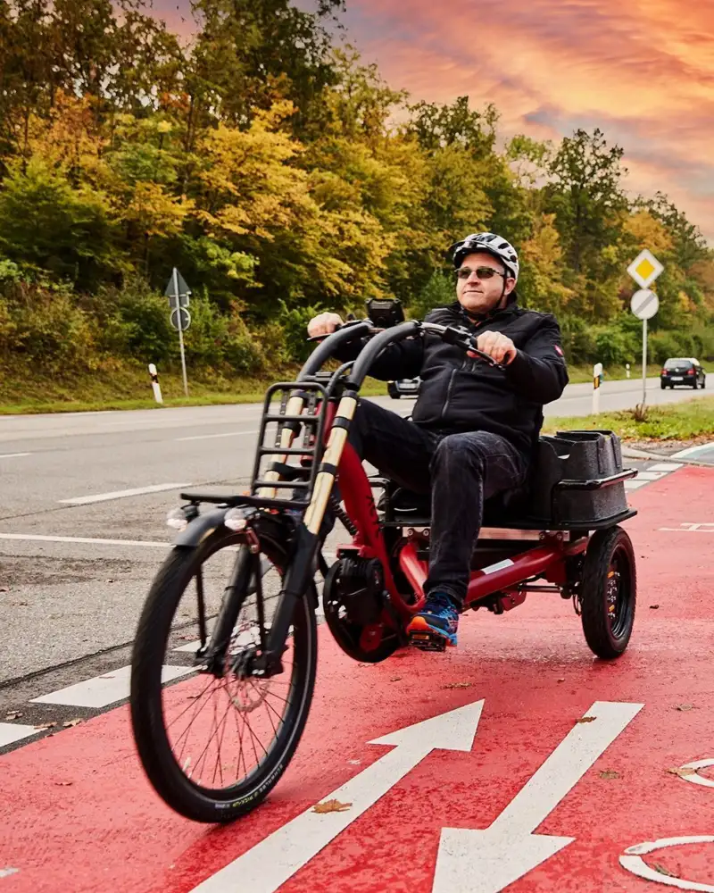 Ein Mann mit Helm und Sonnenbrille ist auf einem Radweg mit einem E-Bike mit Ladefläche unterwegs.
