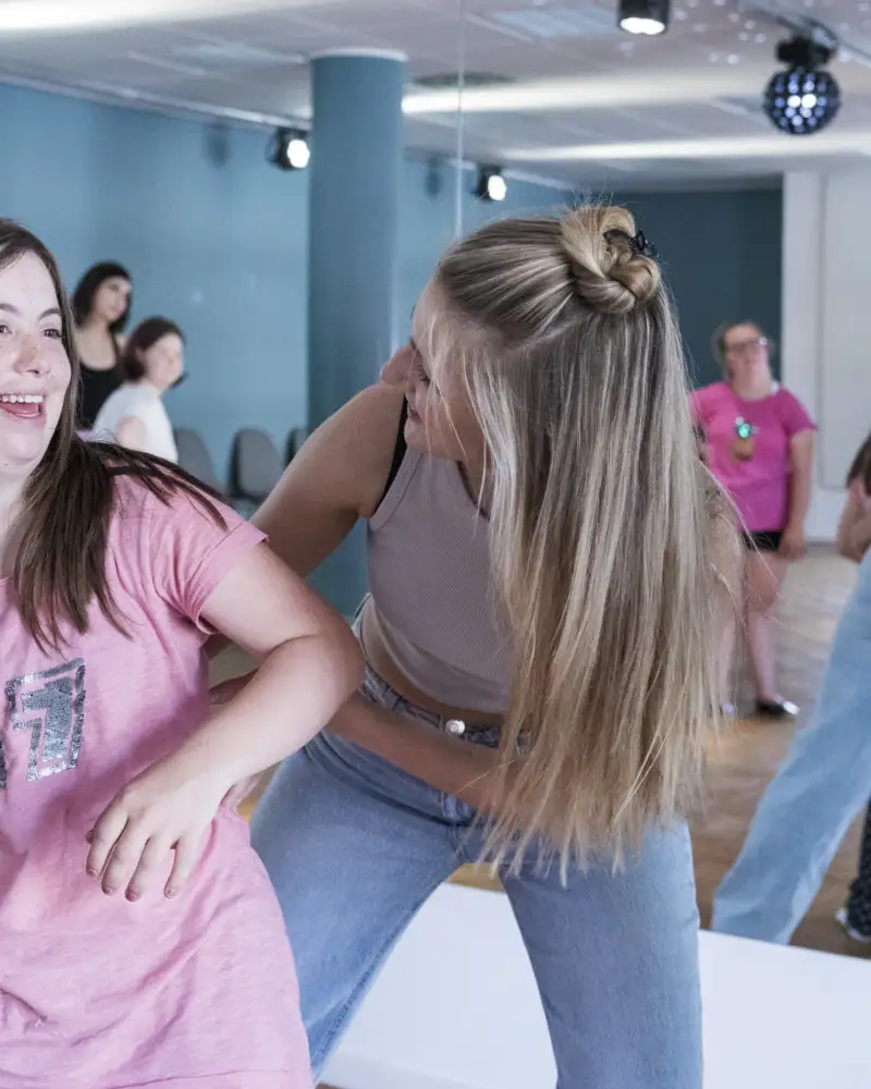 Zwei Mädchen tanzen nebeneinander und schauen sich dabei an. Sie stehen mit dem Rücken vor einem großen Spiegel. Darin sind noch weitere Kinder und Jugendliche zu sehen.