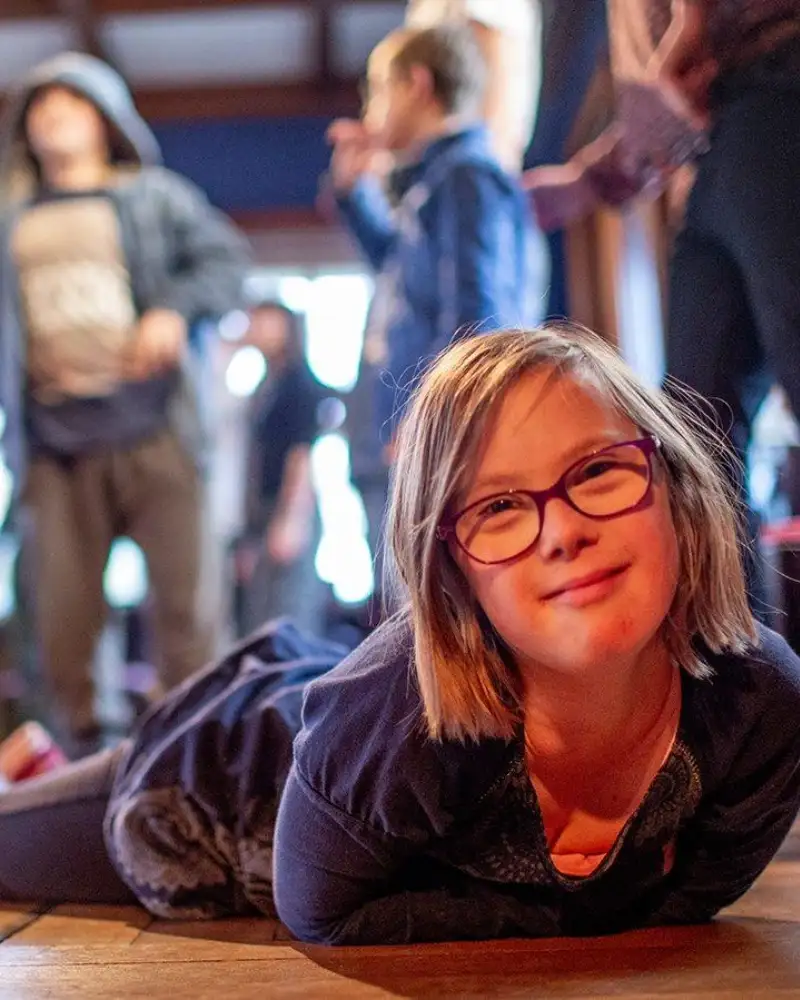 Eine junge Frau mit Downsyndrom liegt auf dem Boden, im Hintergrund steht eine Gruppe von weiteren Personen.