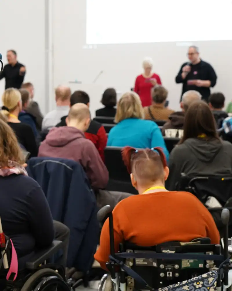 In einem Saal sitzen viele Menschen, darunter auch einige Rollstuhlnutzer*innen. Vor den Zuhörer*innen stehen ein Mann und eine Frau mit Mikrofonen.