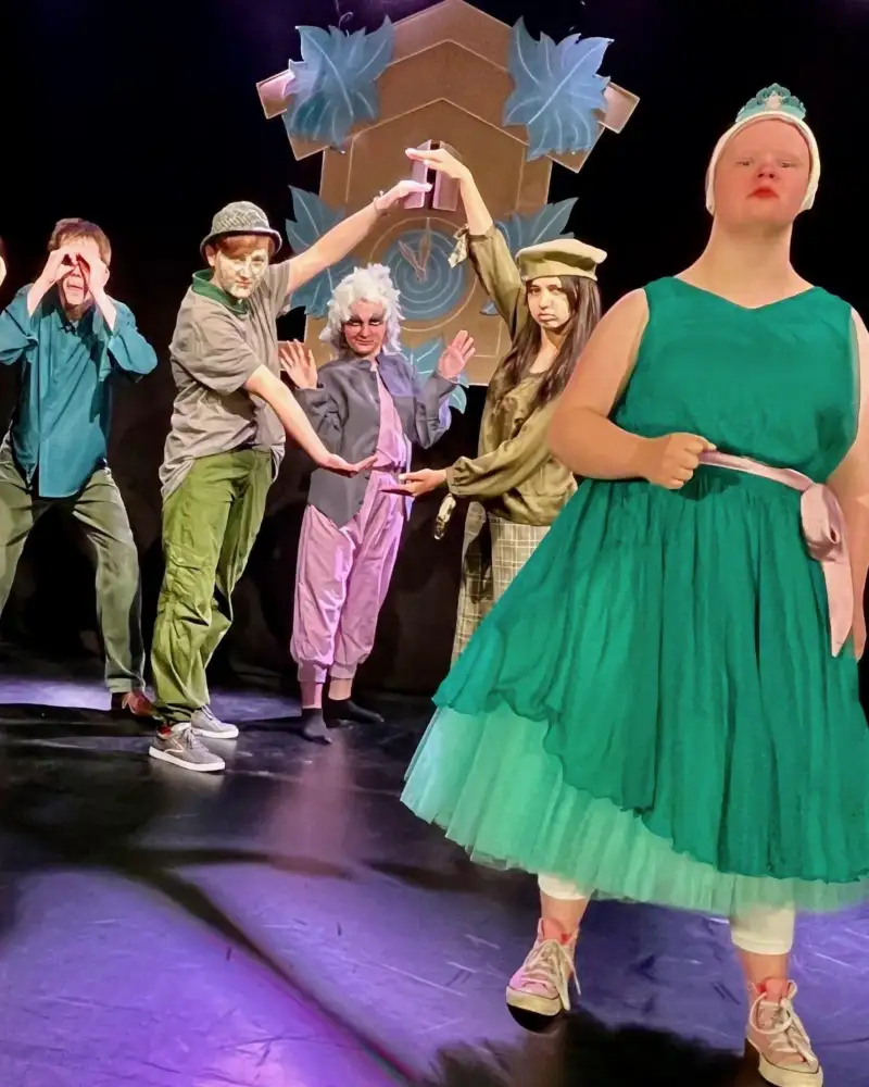 Vorne rechts auf einer Bühne steht eine junge Darstellerin mit Downsyndrom im grünen Petticoat-Kleid. Vorne links steht eine Darstellerin in alpenländischer Kostümierung. Im Hintergrund stehen weitere Darsteller*innen.