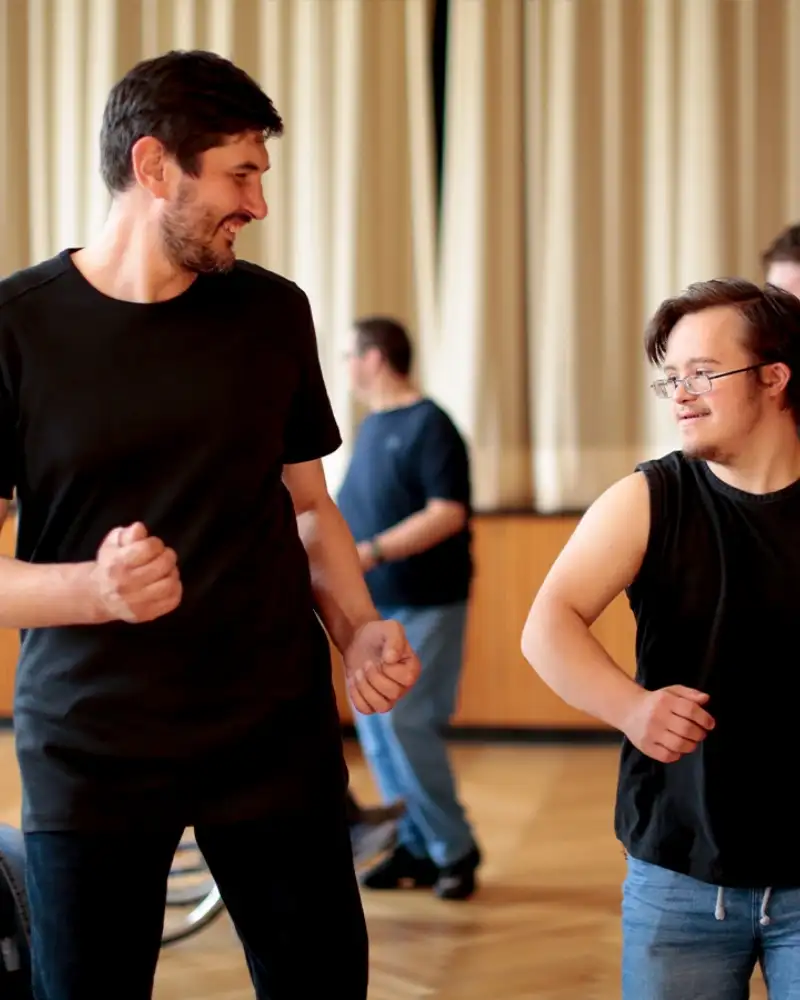 Zwei Männer tanzen nebeneinander. Der kleinere von beiden hat das Downsyndrom. Im Hintergrund tanzen weitere Personen, eine von ihnen sitzt im Rollstuhl.