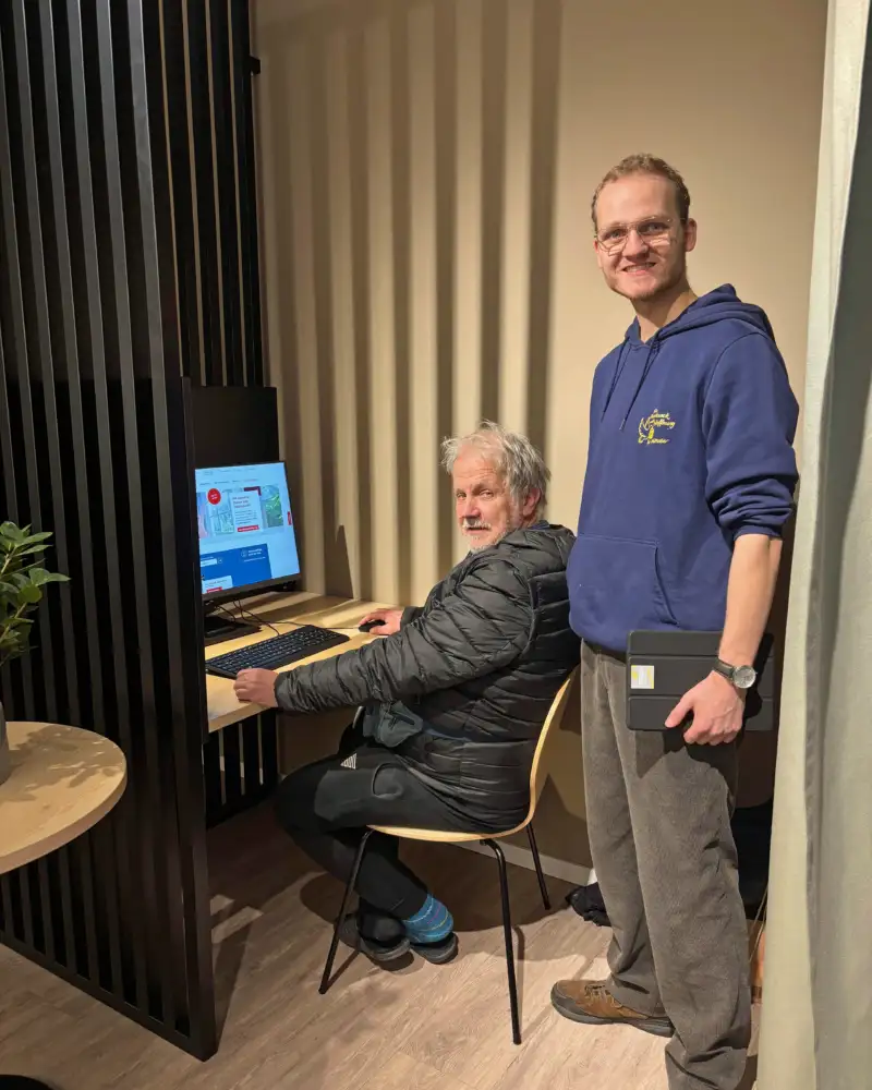 Ein grauhaariger Mann mit Bart sitzt vor einem Computer. Neben ihm steht ein jüngerer Mann im Hoodie.