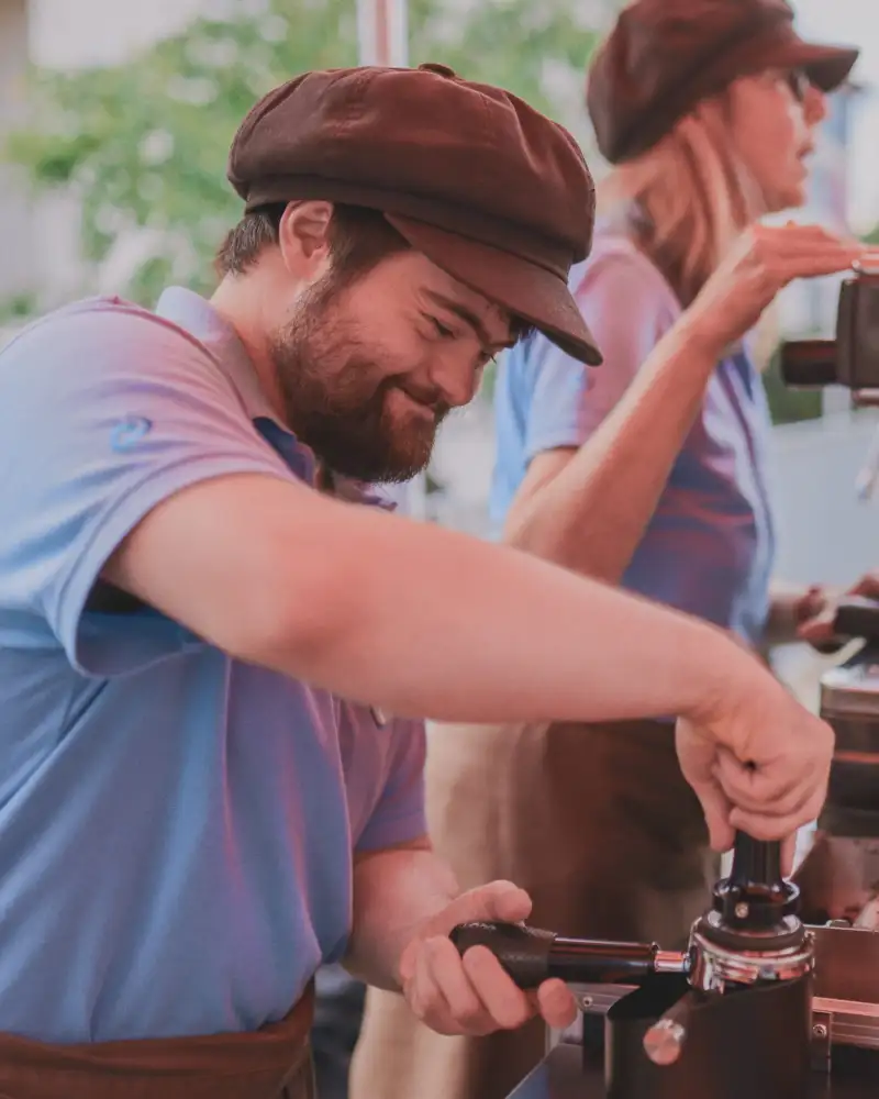 Ein Mann bedient eine Esspressomaschine. Neben ihm steht eine Frau, die eine andere Maschine bedient und mit Kunden spricht.