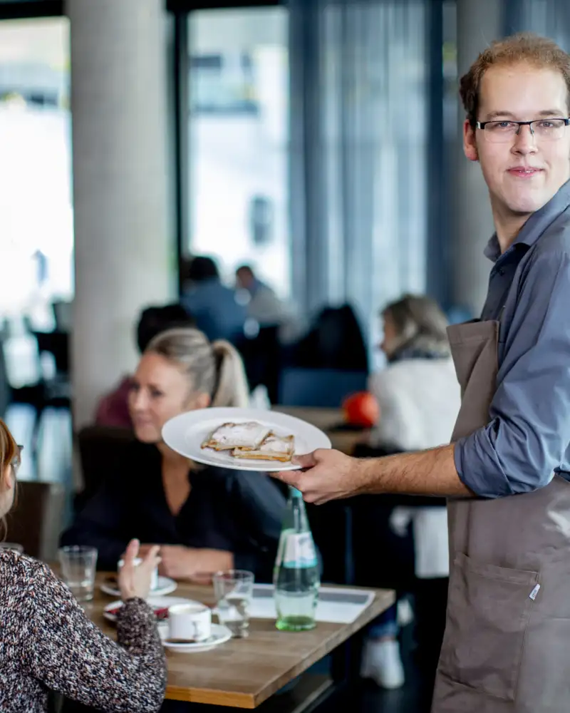 Ein junger Mann mit Schürze serviert Palatschinken an einem Tisch, an dem zwei Frauen sitzen.