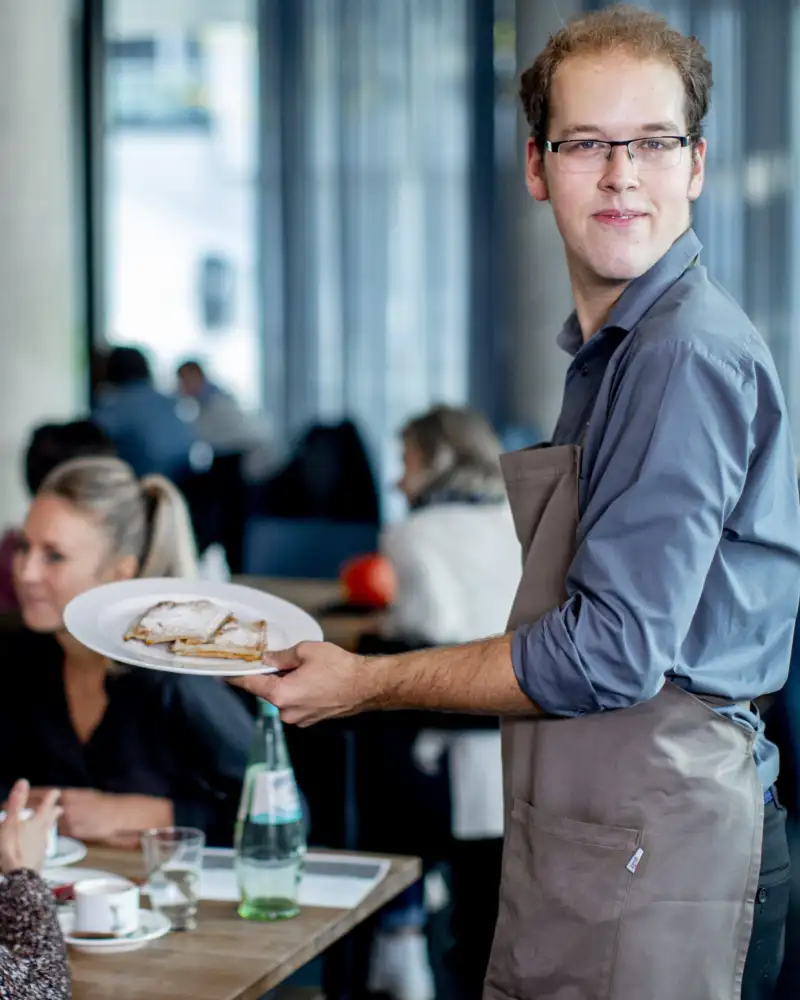 Ein junger Mann mit Schürze serviert Crèpes an einem Tisch, an dem zwei Frauen sitzen.