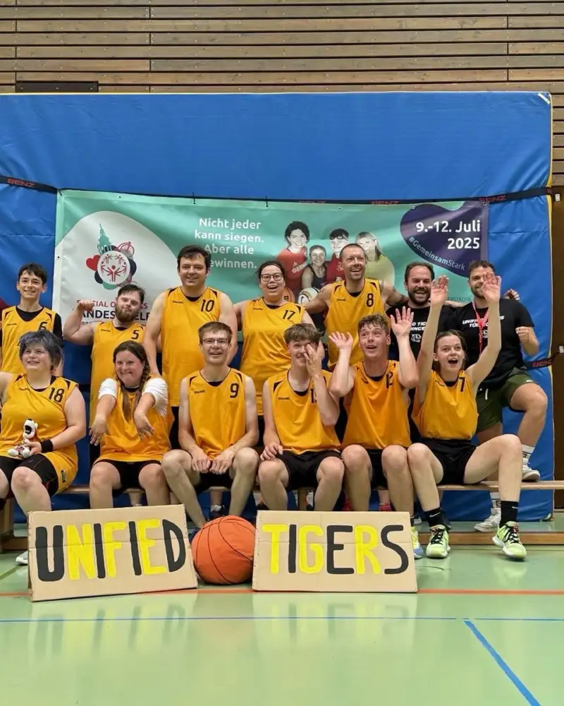 Eine Gruppe von Männern und Frauen unterschiedlichen Alters. Sie befinden sich in einer Turnhalle und tragen gelb-schwarze Sporttrikots. Vor ihnen stehen selbstgemalte Plakate mit der Aufschrift "Unified Tigers".