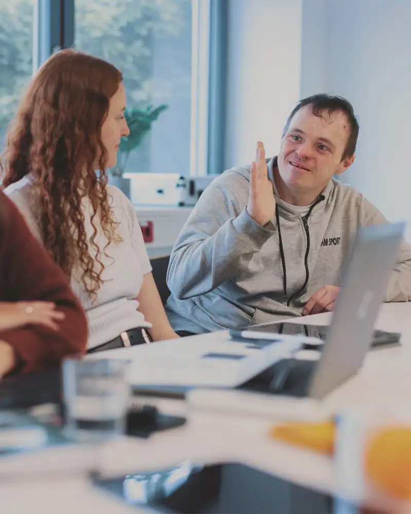 Ein Mann mit Downsyndrom spricht mit zwei Frauen. Die drei sitzen gemeinsam an einem Tisch in einem Besprechungsraum.