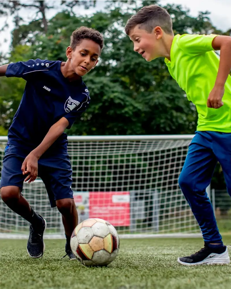 Zwei Jungen kämpfen auf einem Fußballplatz um den Ball. Im Hintergrund ist das Tor zu sehen.