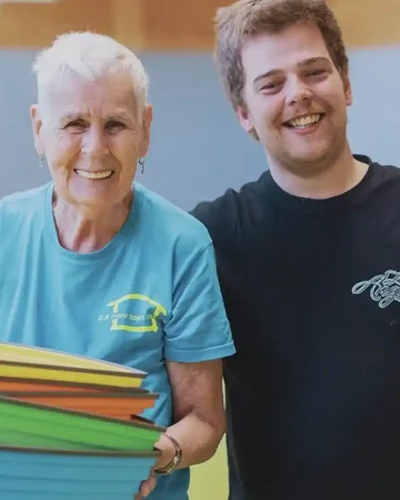 Eine Seniorin mit kurzen weißen Haaren und hellblauem T-Shirt steht in einer Turnhalle neben einem jungen Mann mit kurzen dunkelblonden Haaren in dunklem T-Shirt, der ihr seinen rechten Arm um die Schulter gelegt hat. Die alte Frau hält bunte Plastikhütchen in ihren Händen.