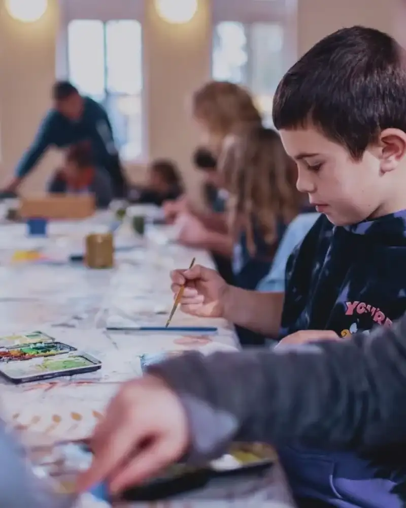 Ein kleiner Junge sitzt mit vielen anderen Kindern an einem langen Tisch und malt.