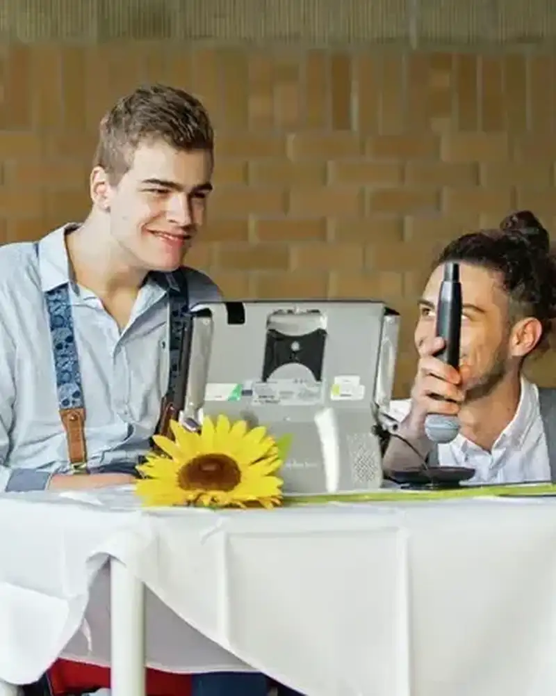 Ein Mann mit geistiger Behinderung sitzt an einem Tisch mit Bildschirm, neben ihm hockt ein anderer Mann.