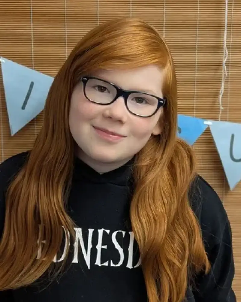 Ein Mädchen mit roten langen Haaren und Brille lächelt in die Kamera.