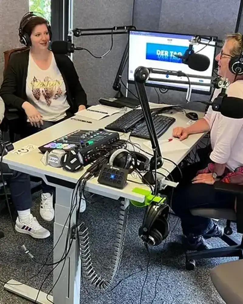 Drei Frauen sitzen an einem Tisch. Sie haben Mikrophone vor dem Gesicht und Tastaturen vor sich liegen. Dahinter ist ein eingeschalteter Monitor angebracht.