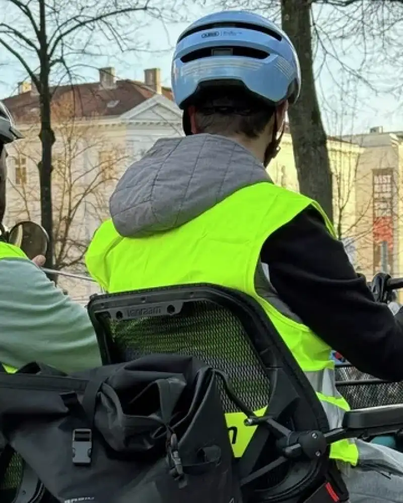 Zwei Personen sind per Rad auf einer Straße unterwegs. Beide tragen Helme und Warnwesten.
