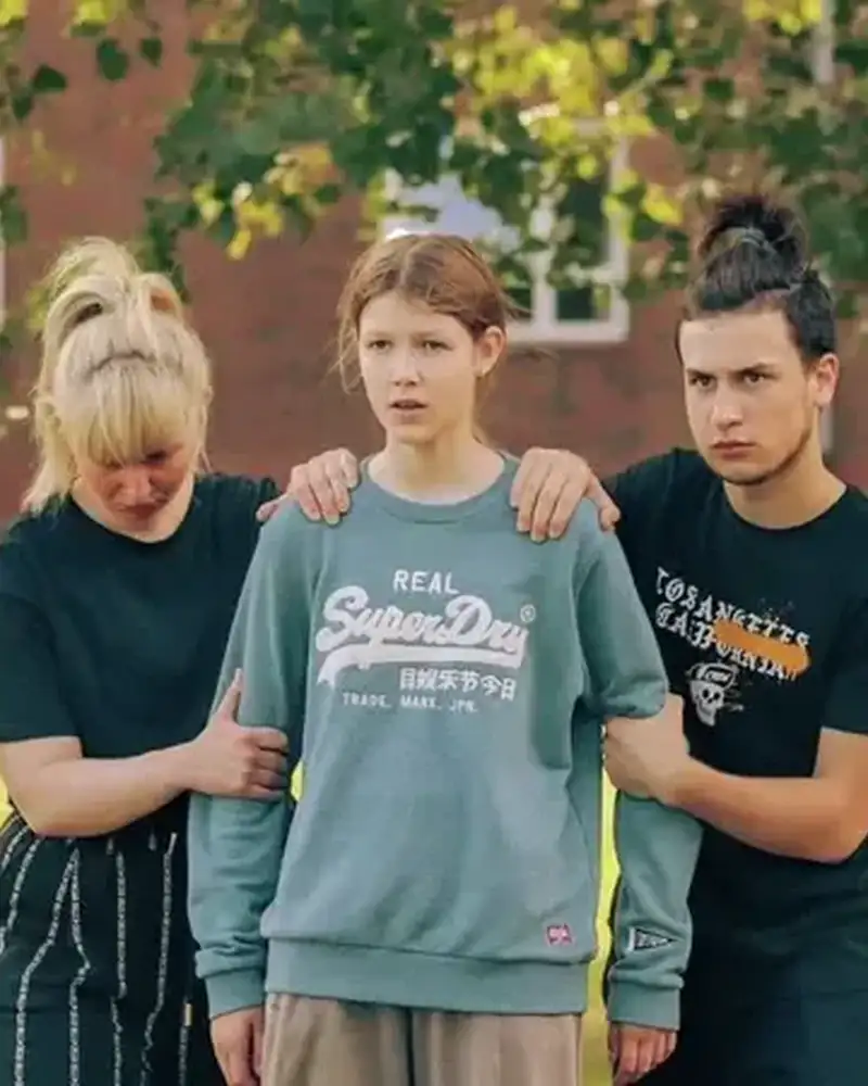 Ein junger Mann und eine junge Frau halten eine Jugendliche an Oberarm und Schulter  unter einem Baum.