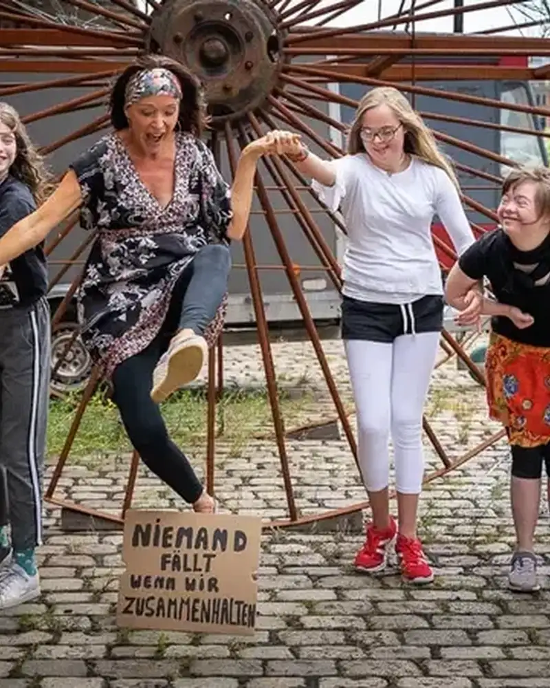 Vier Kinder und Jugendliche mit Behinderung und eine Frau ohne Behinderung stehen in Reihe und halten sich an den Händen. Die Frau springt über ein Schild, auf dem steht: "Niemand fällt wenn wir zusammenhalten".