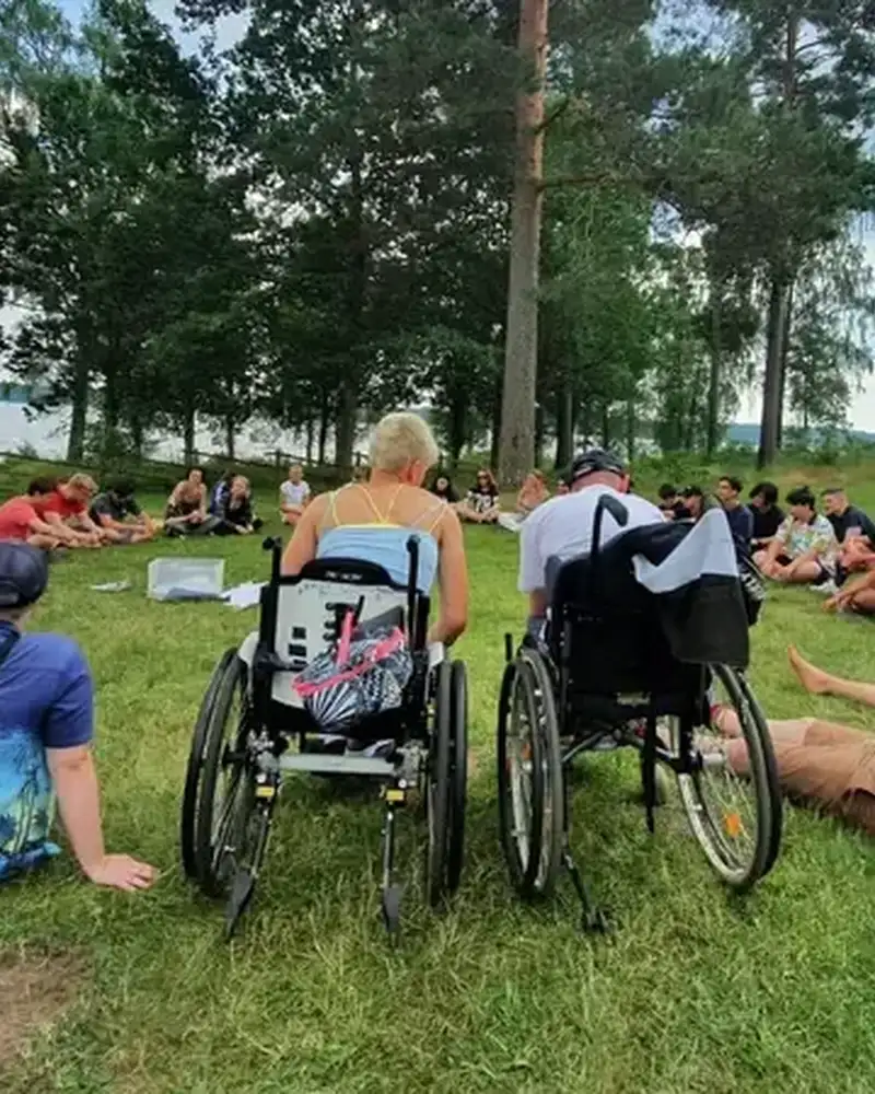 Eine Gruppe Jugendlicher, darunter auch Rollstuhlfahrer*innen, sitzen im Kreis auf einer Wiese