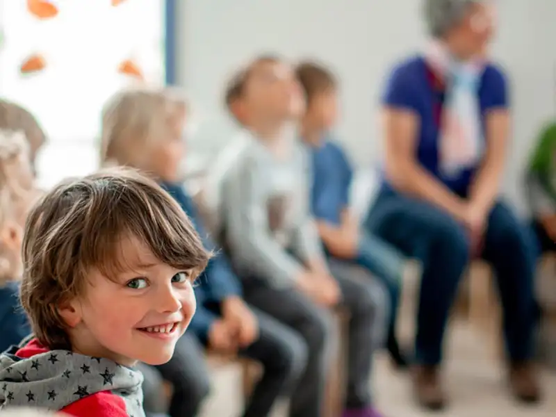 Ein kleiner Junge lacht in die Kamera. Im Hintergrund ist unscharf eine Reihe von Kindern mit einer Mitarbeiterin der Kita zu sehen.