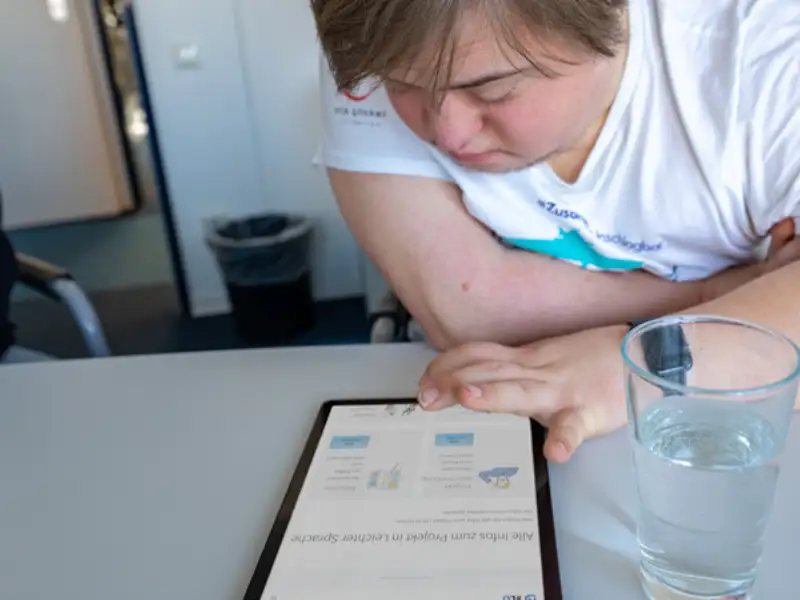 Ein junger Mann mit Downsyndrom tippt auf ein Tablet, das vor ihm liegt.