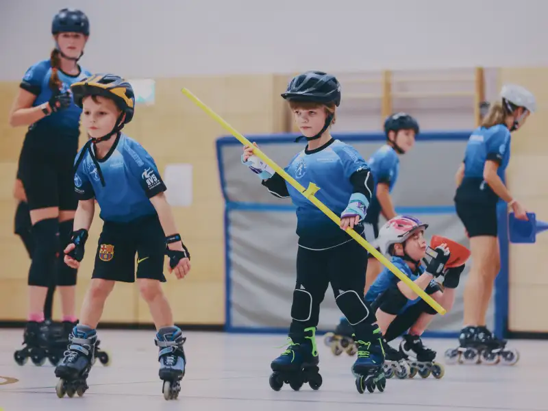 Zwei kleine Jungen mit blauen Trikots und Helmen auf Inlineskates in einer Halle. Einer von ihnen trägt einen langen Stock. Im Hintergrund sind weitere Personen auf Inlineskates.