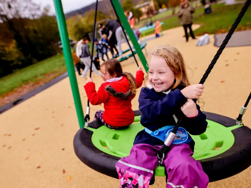 Zwei kleine Mädchen sitzen mit dem Rücken zueinander auf einer Nestschaukel auf einem Spielplatz.