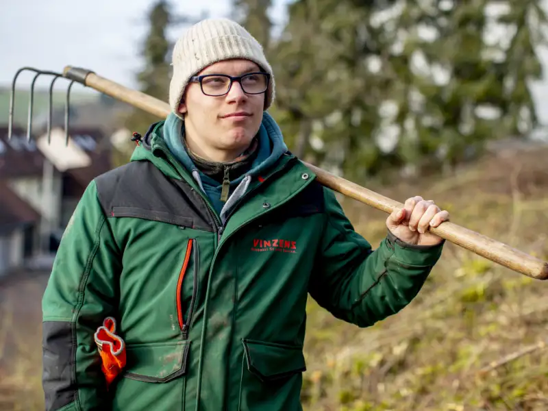 Ein Mann im grünen Anorak trägt eine Harke auf der Schulter.