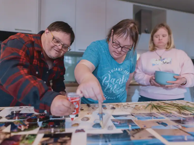 Drei Personen mit Downsyndrom sitzen an einem Tisch, auf dem viele Urlaubsfotos liegen.