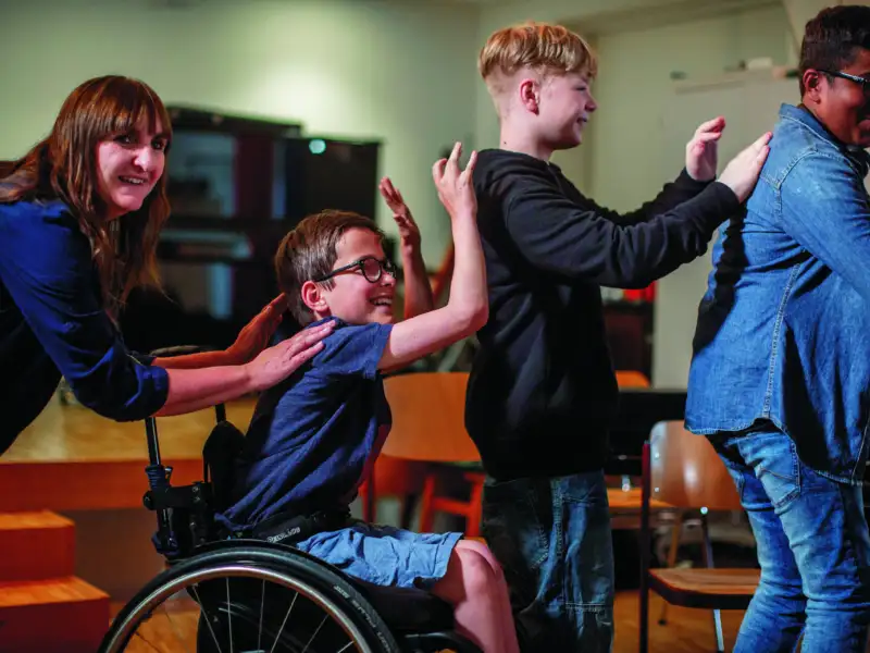Vier Schüler*innen- darunter eine im Rollstuhl - bewegen sich hintereinander. Sie haben ihre Hände auf die Schulter der Person vor ihnen gelegt.