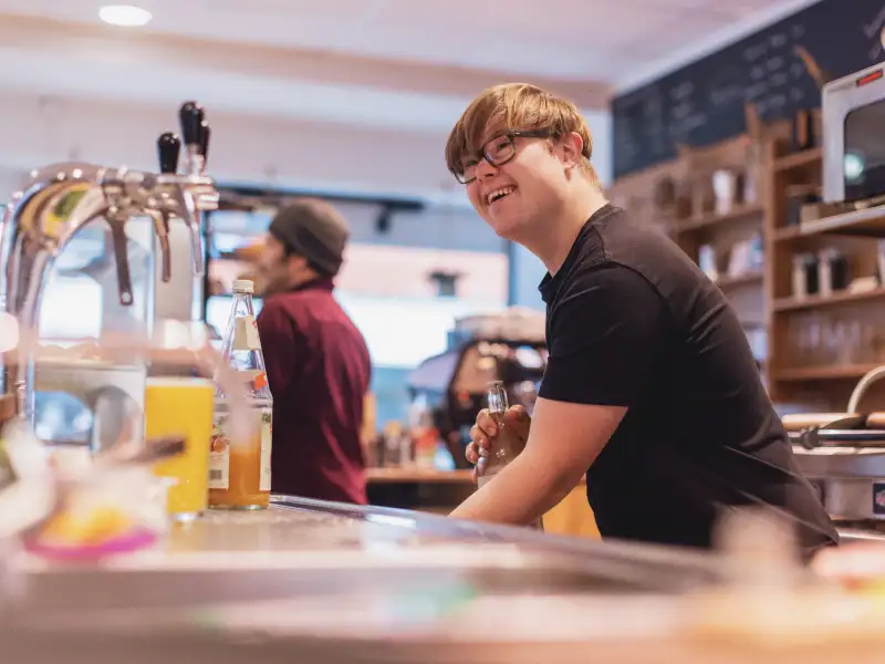 Ein junger Mann mit Downsyndrom arbeitet in einem Bistro hinterm Tresen.