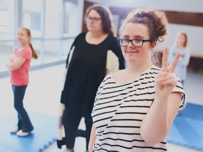 Eine Jugendliche zeigt das Victory-Zeichen mit gespreizten Zeige- und Mittelfinger. Im Hintergrund sthen weitere Personen.