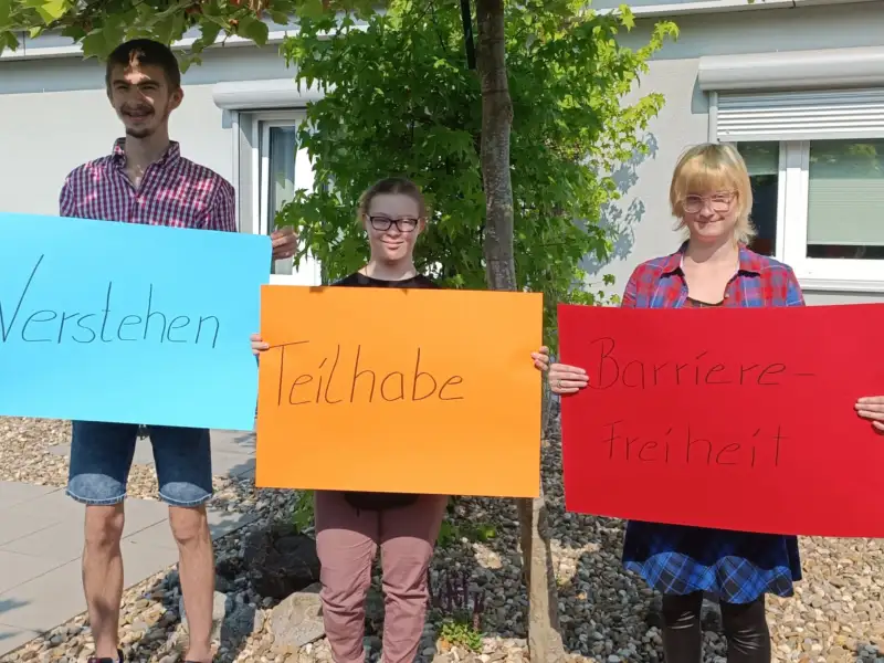 Nebeneinander stehen ein Mann und zwei Frauen, von denen eine das Downsyndrom hat. Jede/r von ihnen trägt ein buntes Plakat mit einem Wort: Verstehen, Teilhabe und Barrierefreiheit.