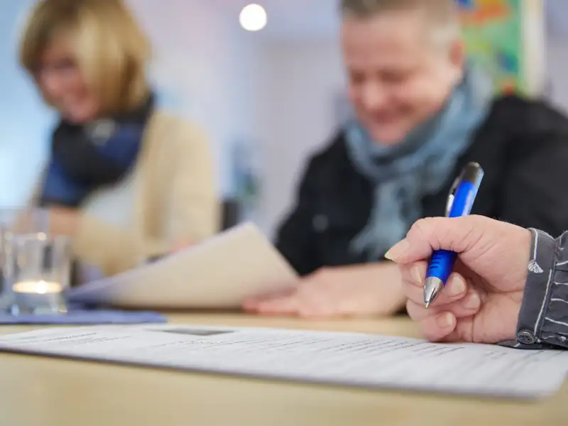 Drei Personen beschäftigen sich mit Formularen, eine dritte, von der man nur die Hand sieht, hält einen Stift über einem Formular.