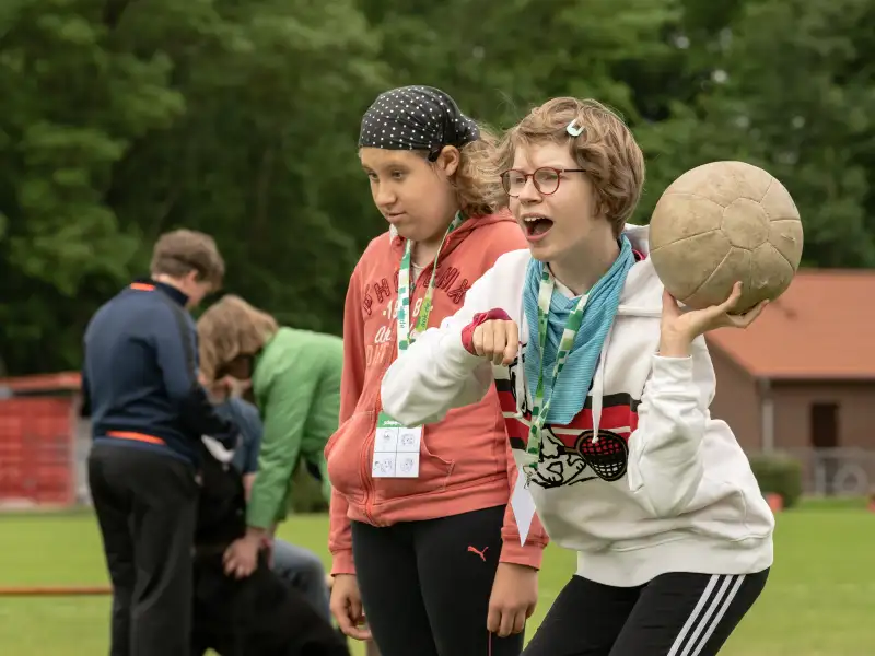 Zwei Mädchen auf einem Sportplatz. Eine von beiden wirft einen Ball.